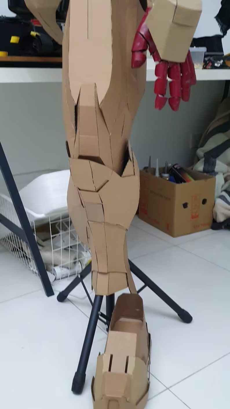 May include: A cardboard model of a robot leg, painted red and brown, with a black stand. The leg is made of multiple cardboard pieces, and the foot is separate.