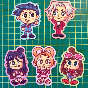 Op de afbeelding: Een verzameling van vijf cartoon karakter stickers. De stickers tonen karakters met grote ogen en verschillende outfits. De karakters hebben verschillende haarkleuren, waaronder blauw, wit en bruin. De stickers staan op een groene rasterachtergrond.