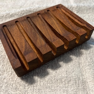 May include: A rectangular wooden soap dish with a rich brown finish. The dish features multiple parallel grooves carved into the surface, designed to hold and drain soap. The wood grain is visible, adding to its natural aesthetic.