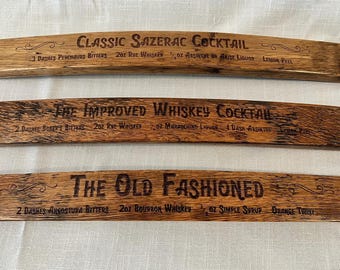 Könnte beinhalten: Drei Holzschilder mit den Namen klassischer Cocktails und ihren Zutaten. Die Schilder sind aus recyceltem Holz gefertigt und haben einen rustikalen Look. Die Cocktails sind der Classic Sazerac, der Improved Whiskey Cocktail und der Old Fashioned.
