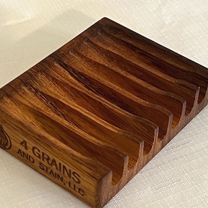 May include: A rectangular wooden soap dish with a grooved design. The dish is made of dark brown wood and features the text "4 GRAINS AND STAIN, LLC" on the side. The dish has a natural wood grain pattern.