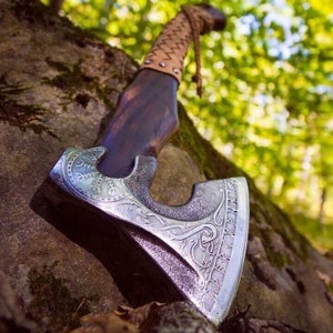 Könnte beinhalten: Eine handgefertigte Axt mit einem dunklen Holzgriff und einem silberfarbenen Kopf. Der Kopf weist aufwendige eingravierte Designs auf. Der Griff ist mit braunem Leder umwickelt. Die Axt lehnt an einem Felsen mit verschwommenem grünem Hintergrund.