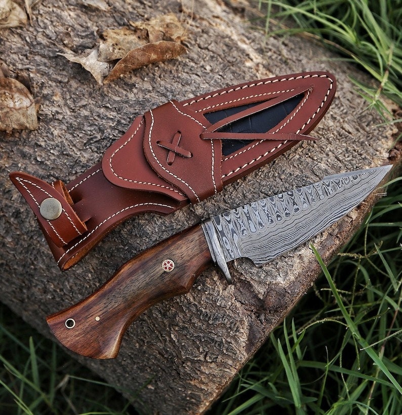 K&ouml;nnte beinhalten: Ein braunes Lederscheide mit einem braunen Holzgriffmesser mit einer gemusterten Klinge. Das Messer liegt auf einer Holzoberfl&auml;che mit gr&uuml;nem Gras im Hintergrund.