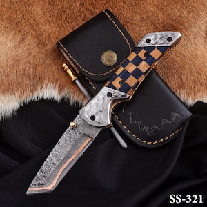 May include: A Damascus steel folding knife with a checkered blue and tan wood handle. The knife has a copper-colored edge and is accompanied by a black leather sheath and a sharpening rod. The text "SS-321" is visible.