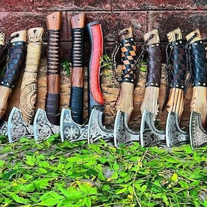 May include: A collection of 15 hand-crafted axes with wooden handles and leather wrappings. The axes have intricate designs on the blades and are displayed against a brick wall.