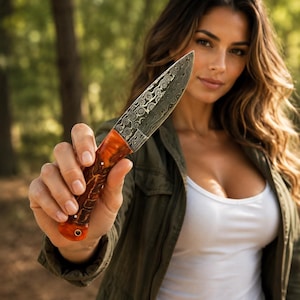 Puede incluir: Un cuchillo de hoja fija con una distintiva hoja de acero de Damasco con un patrón complejo y arremolinado, y un mango de resina naranja translúcida pulida con texturas internas visibles y un pequeño orificio metálico en la base.