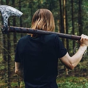 May include: A large axe with a wide, curved blade and a long wooden handle. The axe head is silver, and the handle is wrapped in dark brown leather. The axe is being held over the shoulder against a backdrop of a forest.