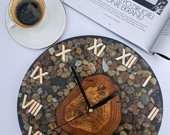 May include: A round clock with Roman numeral markings on a black background. The clock face is filled with small, colorful stones embedded in clear resin. The clock hands are black and gold. The clock is set against a white background.