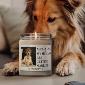 May include: A lit soy wax candle in a glass jar with a silver lid. The label features a photo of a puppy and the text "Smells Like My Humans Are Getting Married" with a wedding date. A dog is in the background.