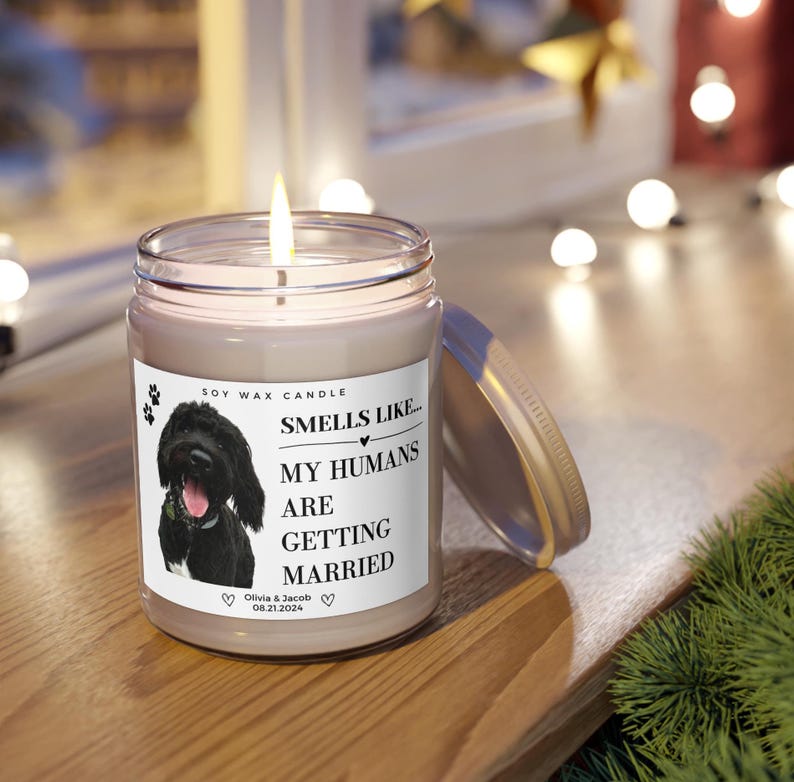 May include: A lit soy wax candle in a clear glass jar with a metal lid. The label features a black dog image and text that reads "Smells Like... My Humans Are Getting Married" with a wedding date. The candle sits on a wooden surface.