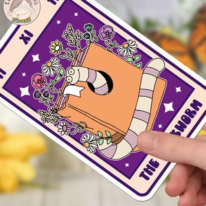 Puede incluir: Una pegatina morada y naranja con un gusano de libro y flores. El texto de la pegatina dice "The Bookworm".