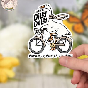 Pu&ograve; includere: Un'oca bianca con una sciarpa va in bicicletta con un cestino. L'oca indossa una sciarpa e ha il testo "Born to Dilly Dally" sul petto. Il testo "Forced to Pick Up the Pace" &egrave; sotto la bicicletta.