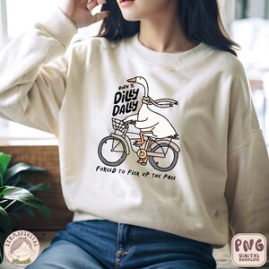 Pu&ograve; includere: Una felpa bianca con una grafica di un'oca che guida una bicicletta. L'oca indossa una sciarpa e il testo sulla felpa recita "Born to Dilly Dally, Forced to Pick Up the Page".
