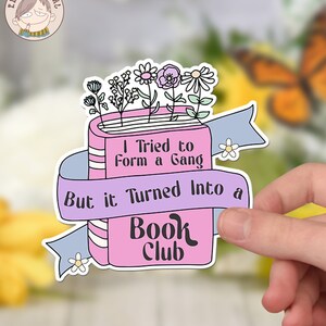 Puede incluir: Pegatina rosa y blanca con una ilustraci&oacute;n de un libro y el texto "Intent&eacute; formar una pandilla, pero se convirti&oacute; en un club de lectura". El libro tiene flores que le salen y una cinta azul alrededor.