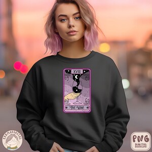 Pu&ograve; includere: Felpa nera con un design di carta dei tarocchi rosa e viola che presenta un bicchiere di vino con una luna crescente e stelle. La carta recita "XVIII Il Vino".