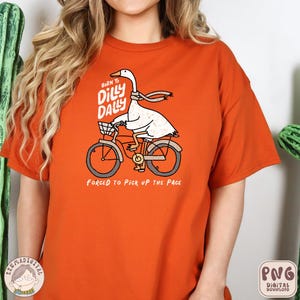Pu&ograve; includere: Maglietta arancione con un'oca bianca che guida una bicicletta. L'oca indossa una sciarpa e il testo sulla maglietta recita "Born to Dilly Dally, Forged to Pick Up the Page".