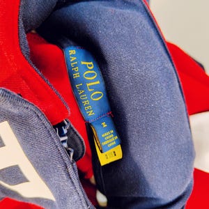 May include: Close-up of a red, white, and navy blue Polo Ralph Lauren jacket. The navy blue collar and label are visible, with the brand name in gold lettering. The jacket is size M and made in China.