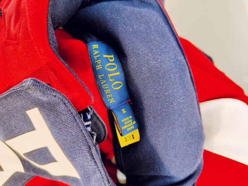May include: Close-up of a red, white, and navy blue Polo Ralph Lauren jacket. The navy blue collar and label are visible, with the brand name in gold lettering. The jacket is size M and made in China.