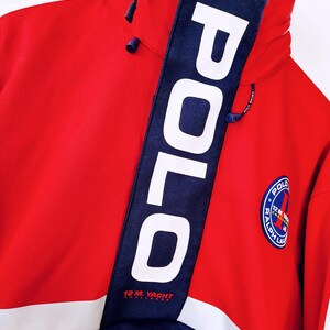 May include: Red and navy blue Polo Sport jacket with a high collar and a zipper. The jacket features a white stripe across the chest and the word "POLO" in large white letters on a navy blue band. A circular patch with text is on the right side.