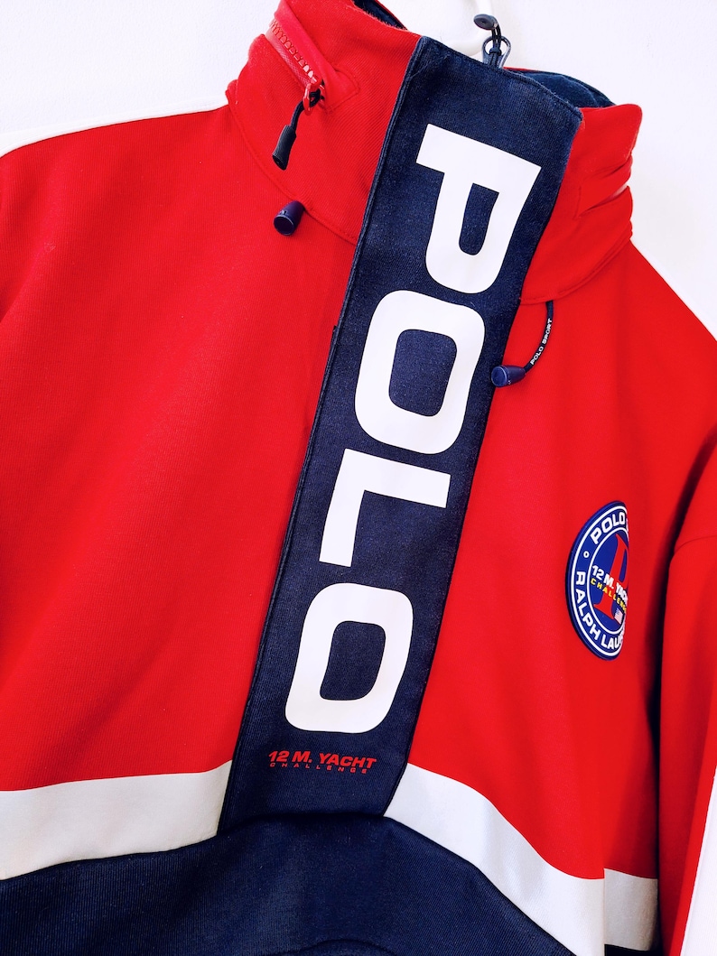 May include: Red and navy blue Polo Sport jacket with a high collar and a zipper. The jacket features a white stripe across the chest and the word "POLO" in large white letters on a navy blue band. A circular patch with text is on the right side.