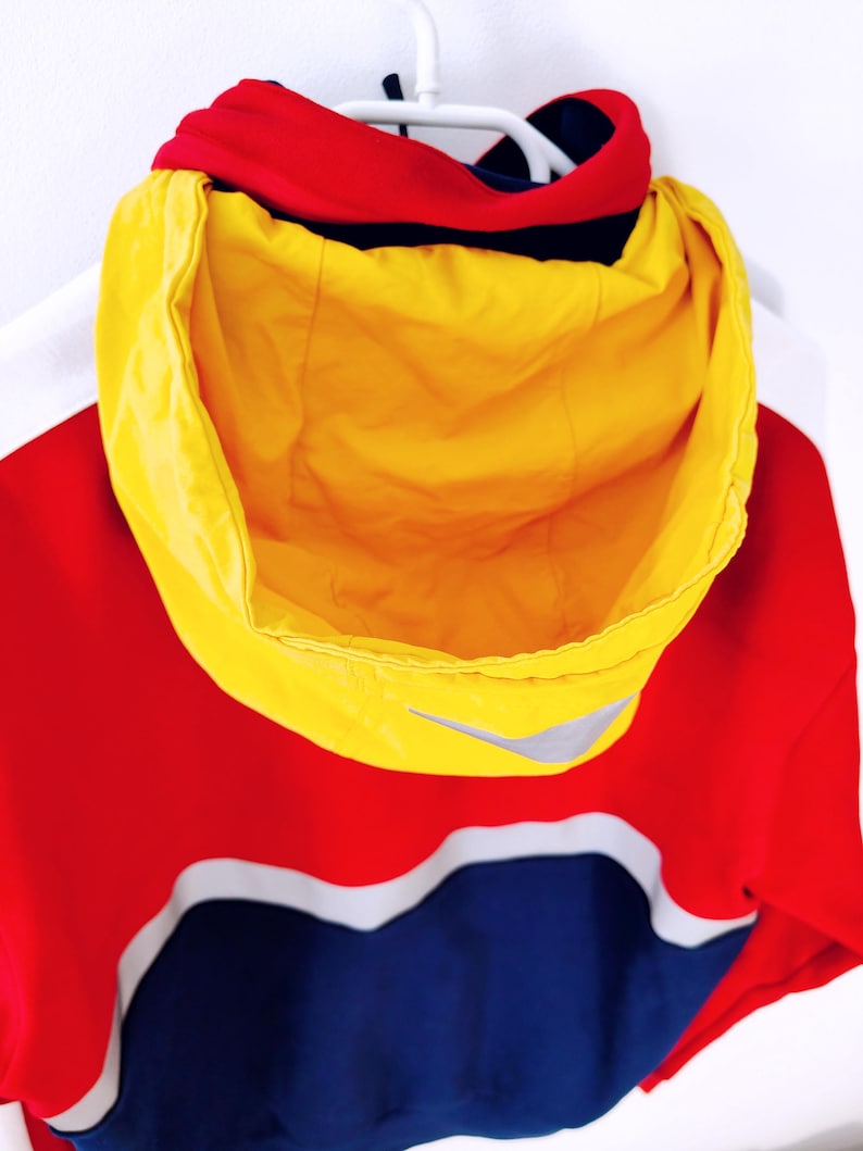May include: A hooded jacket in red, yellow, and blue, with a white accent stripe. The hood is bright yellow, and the jacket features a small silver logo. The jacket is hanging on a white hanger.