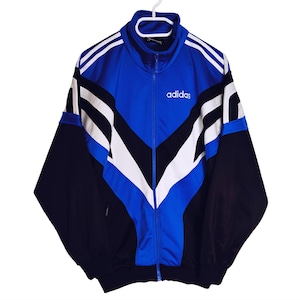 Puede incluir: Chaqueta deportiva Adidas azul, negra y blanca con cierre de cremallera completo. La chaqueta presenta el logo de Adidas en blanco en el pecho y rayas blancas en las mangas. La chaqueta está colgada de una percha blanca.