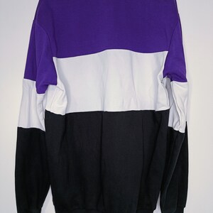 Puede incluir: Una sudadera con bloques de color en morado, blanco y negro. La sudadera est&aacute; colgada de una percha blanca sobre un fondo blanco. El dise&ntilde;o presenta un patr&oacute;n de bloques de color horizontales con los colores dispuestos de arriba a abajo.