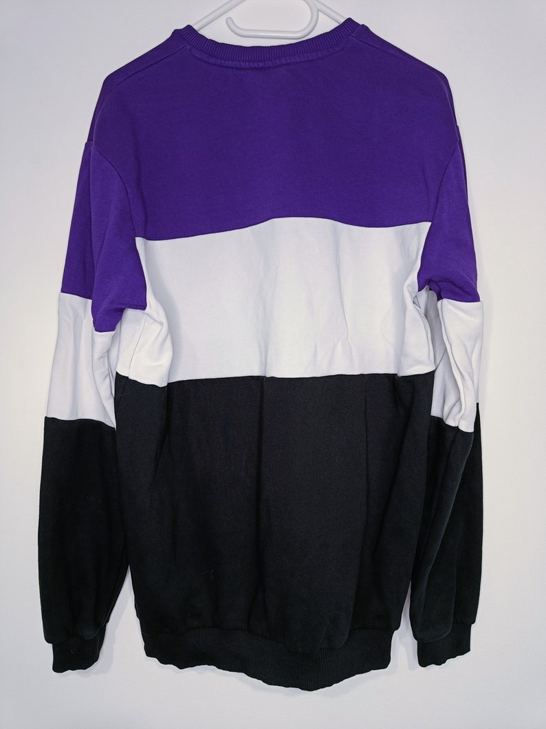 Puede incluir: Una sudadera con bloques de color en morado, blanco y negro. La sudadera est&aacute; colgada de una percha blanca sobre un fondo blanco. El dise&ntilde;o presenta un patr&oacute;n de bloques de color horizontales con los colores dispuestos de arriba a abajo.