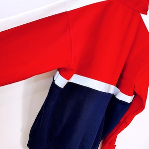 May include: A color-blocked jacket in red, white, and navy blue. The jacket features a high collar and a full-length zipper. The sleeves are long, and the jacket appears to be made of a soft, comfortable material. The design is sporty and casual.