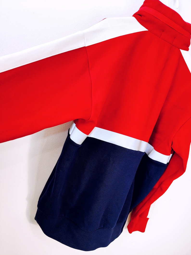 May include: A color-blocked jacket in red, white, and navy blue. The jacket features a high collar and a full-length zipper. The sleeves are long, and the jacket appears to be made of a soft, comfortable material. The design is sporty and casual.