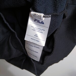 Puede incluir: Primer plano de la etiqueta de una sudadera FILA azul marino. La etiqueta muestra el logotipo de FILA, los datos de la empresa y la composici&oacute;n de la tela. La sudadera est&aacute; fabricada en Turqu&iacute;a y est&aacute; compuesta de algod&oacute;n y poli&eacute;ster.