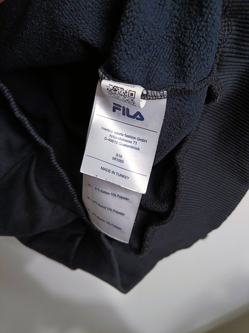 Puede incluir: Primer plano de la etiqueta de una sudadera FILA azul marino. La etiqueta muestra el logotipo de FILA, los datos de la empresa y la composici&oacute;n de la tela. La sudadera est&aacute; fabricada en Turqu&iacute;a y est&aacute; compuesta de algod&oacute;n y poli&eacute;ster.