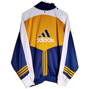 Puede incluir: Chaqueta deportiva Adidas vintage con diseño de bloques de color. La chaqueta presenta una combinación de colores azul marino, blanco y dorado con el logo de Adidas en el pecho. Tiene cuello alto y puños acanalados.