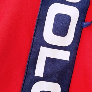 May include: Close-up of a red garment with a navy blue stripe. The stripe features the word "POLO" in large white capital letters. A blue drawstring is visible at the top. The fabric appears to be a knit material.