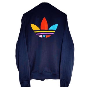 Puede incluir: Chaqueta deportiva azul marino con cierre de cremallera completa. La parte trasera presenta un gran logotipo colorido con detalles en naranja, rojo, amarillo, rosa y azul. La chaqueta tiene cuello alto y rayas verticales en las mangas.