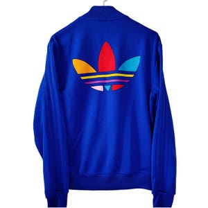 May include: A royal blue track jacket with a full-zip closure and a stand-up collar. The back features a large, colorful logo with orange, yellow, red, pink, purple, and teal accents. The jacket is hanging on a white hanger.