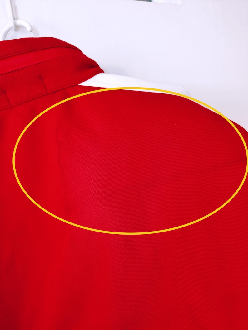 May include: A close-up of a red collared shirt hanging on a white hanger. The shirt is a solid, vibrant red color, with a smooth, slightly shiny texture. The collar is visible, and the shirt appears to be made of a lightweight fabric.