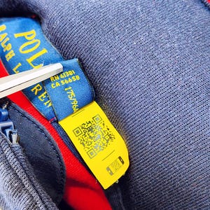 May include: Close-up of a navy blue garment with a red interior. A blue tag with the text "POLO RALPH LAUREN" is visible, along with a yellow tag featuring a QR code. Scissors are partially visible.