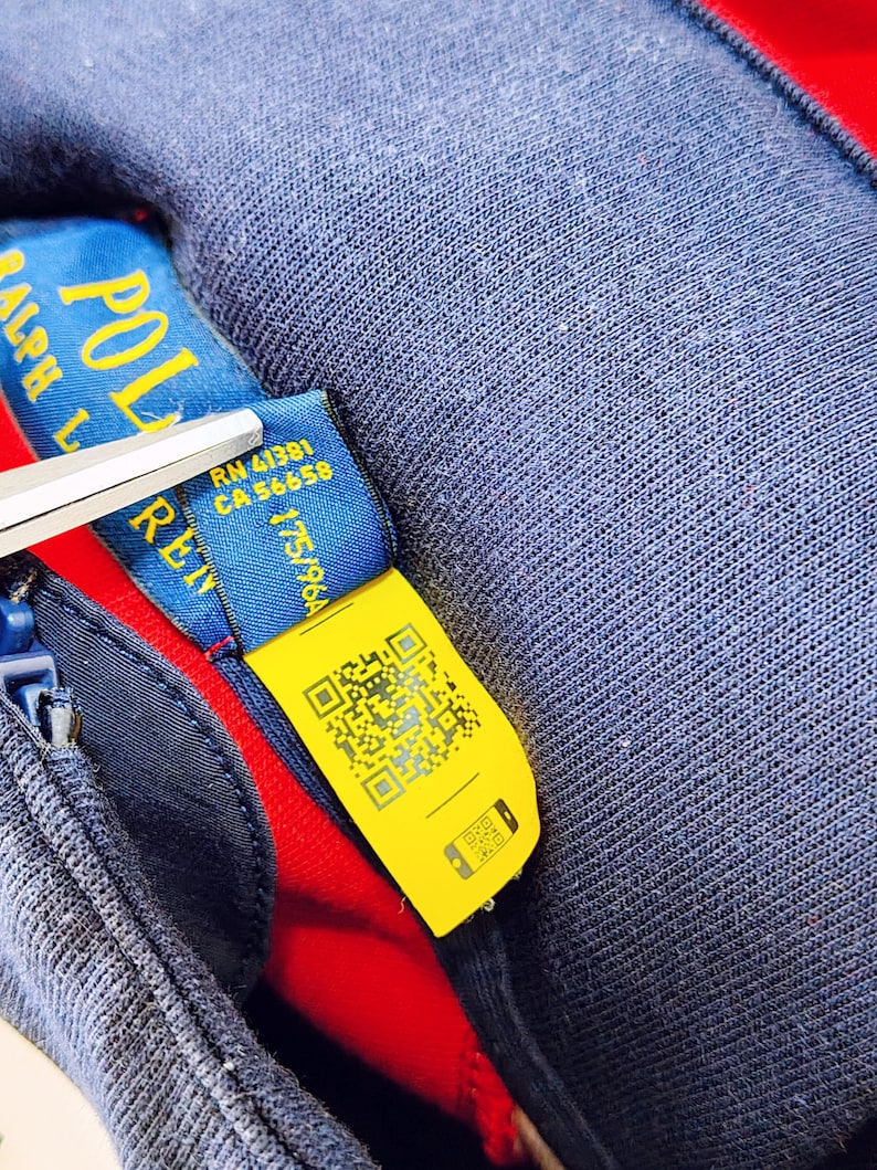 May include: Close-up of a navy blue garment with a red interior. A blue tag with the text "POLO RALPH LAUREN" is visible, along with a yellow tag featuring a QR code. Scissors are partially visible.