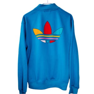 May include: A bright blue zip-up track jacket with a colorful logo on the back. The logo features a trefoil design with red, yellow, orange, pink, and teal accents. The jacket has a high collar and vertical stripes on the sleeves.