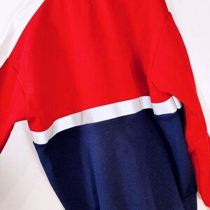 May include: A color-block sweatshirt in red, white, and navy blue. The top section is red with white accents on the shoulders and sleeves. The bottom section is navy blue, creating a bold, sporty look. The sweatshirt has a high collar.