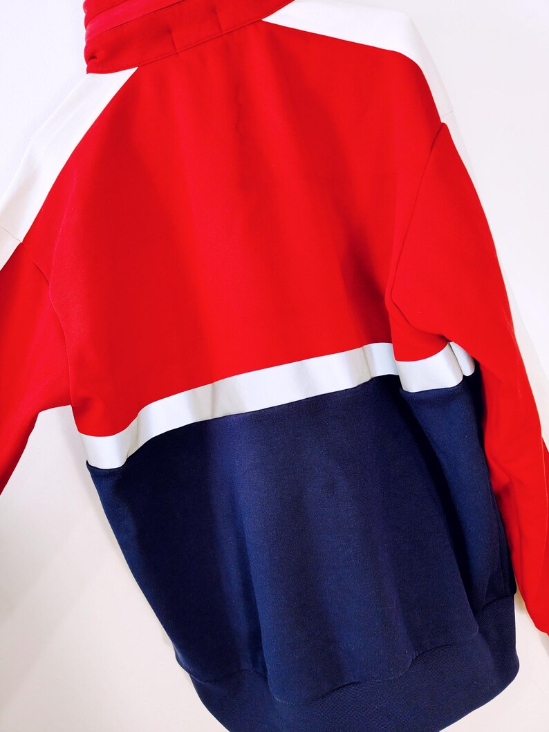 May include: A color-block sweatshirt in red, white, and navy blue. The top section is red with white accents on the shoulders and sleeves. The bottom section is navy blue, creating a bold, sporty look. The sweatshirt has a high collar.