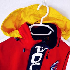 May include: A red and yellow hooded jacket with a navy blue collar and accents. The jacket features the word "POLO" in large white letters on the collar and a circular patch with text. The jacket is hanging on a white hanger.
