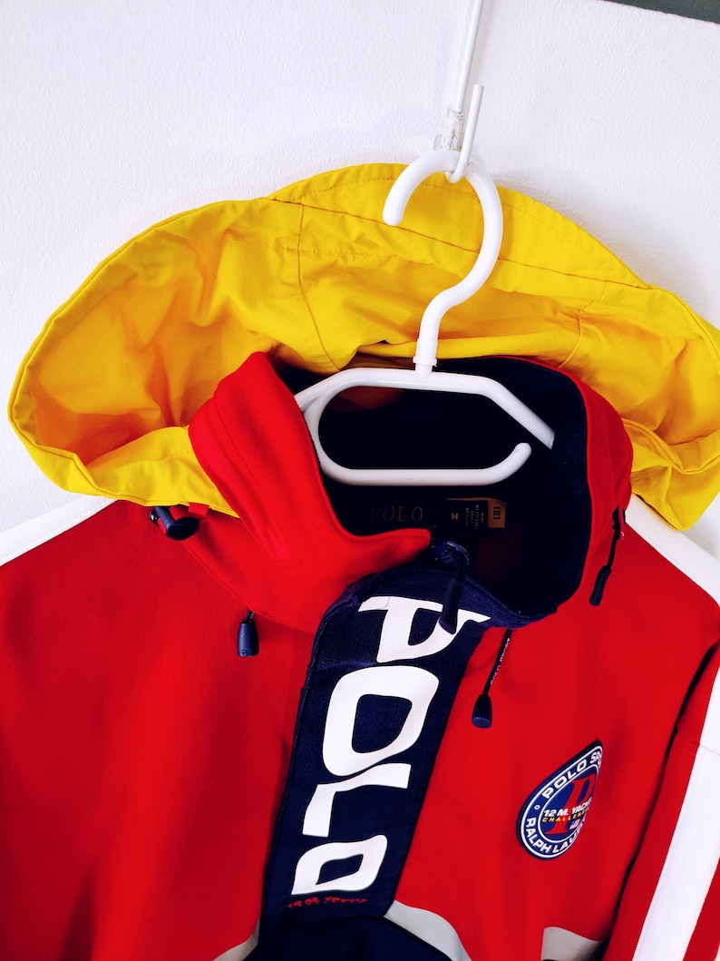 May include: A red and yellow hooded jacket with a navy blue collar and accents. The jacket features the word "POLO" in large white letters on the collar and a circular patch with text. The jacket is hanging on a white hanger.