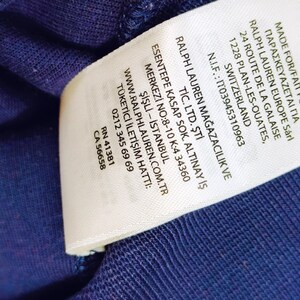 May include: Close-up of a white clothing tag with black text. The tag includes the text "MADE FOR/FAIT POUR/" and "RALPH LAUREN EUROPE S&aacute;rl". Additional text includes a website address and a phone number.