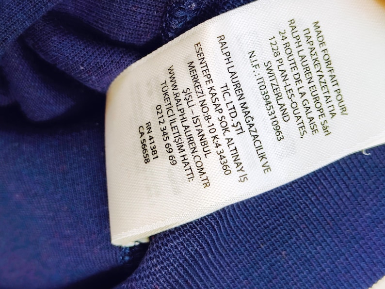May include: Close-up of a white clothing tag with black text. The tag includes the text "MADE FOR/FAIT POUR/" and "RALPH LAUREN EUROPE S&aacute;rl". Additional text includes a website address and a phone number.