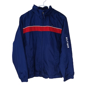 Op de afbeelding: Een marineblauwe jas met een rode streep over de borst. De jas heeft een ritssluiting en een klein logo op de borst. Het woord "HILFIGER" staat verticaal op de mouw gedrukt. De jas hangt aan een witte hanger.