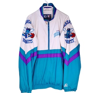 May include: A vintage Charlotte Hornets Starter jacket in white, teal, and purple. The jacket features the Hornets logo on the sleeves and chest, with a zip-up front and a stand-up collar. The jacket is hanging on a white hanger.