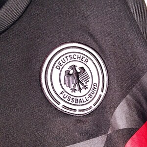 Może przedstawiać: Zbliżenie czarnej koszulki sportowej z emblematem niemieckiej drużyny piłkarskiej. Emblemat to białe koło z napisem "DEUTSCHER FUSSBALL-BUND" i czarnym orłem. Koszulka ma czarno-szary geometryczny wz&oacute;r.