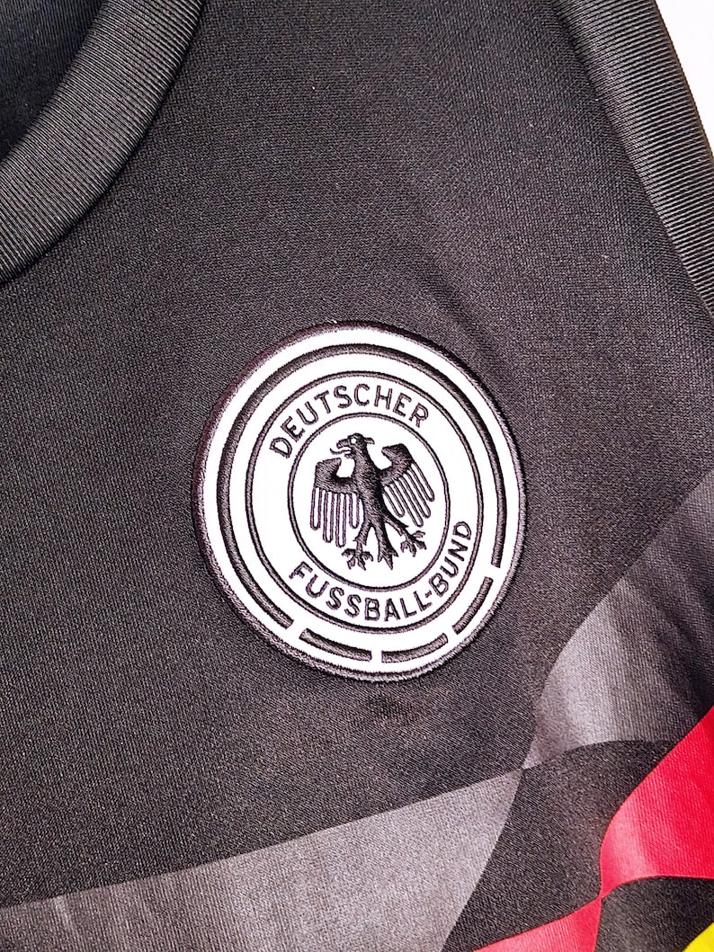 Może przedstawiać: Zbliżenie czarnej koszulki sportowej z emblematem niemieckiej drużyny piłkarskiej. Emblemat to białe koło z napisem "DEUTSCHER FUSSBALL-BUND" i czarnym orłem. Koszulka ma czarno-szary geometryczny wz&oacute;r.